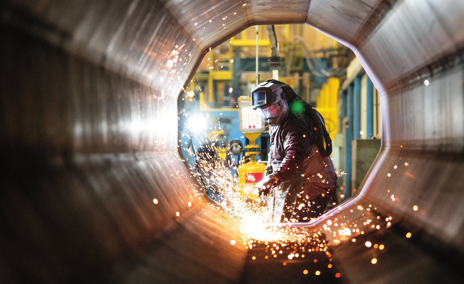 A welder working inside a large steel pipe, sparks flying